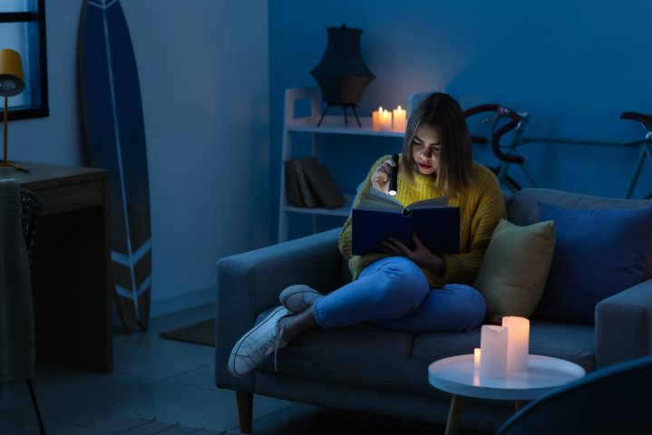 Young,Woman,Reading,Book,With,Flashlight,At,Home,During,Blackout
